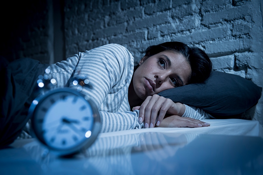 A woman lying in bed at night, unable to sleep, with a distressed expression on her face. An alarm clock on the bedside table shows the time.