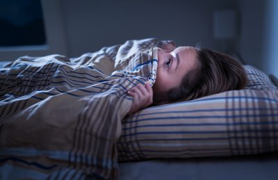 A woman lying in bed with a frightened expression, partially covered by a blanket, indicating fear or distress during sleep.