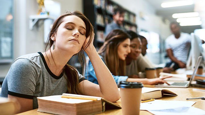 A young woman falling asleep while studying in a library, surrounded by other students.